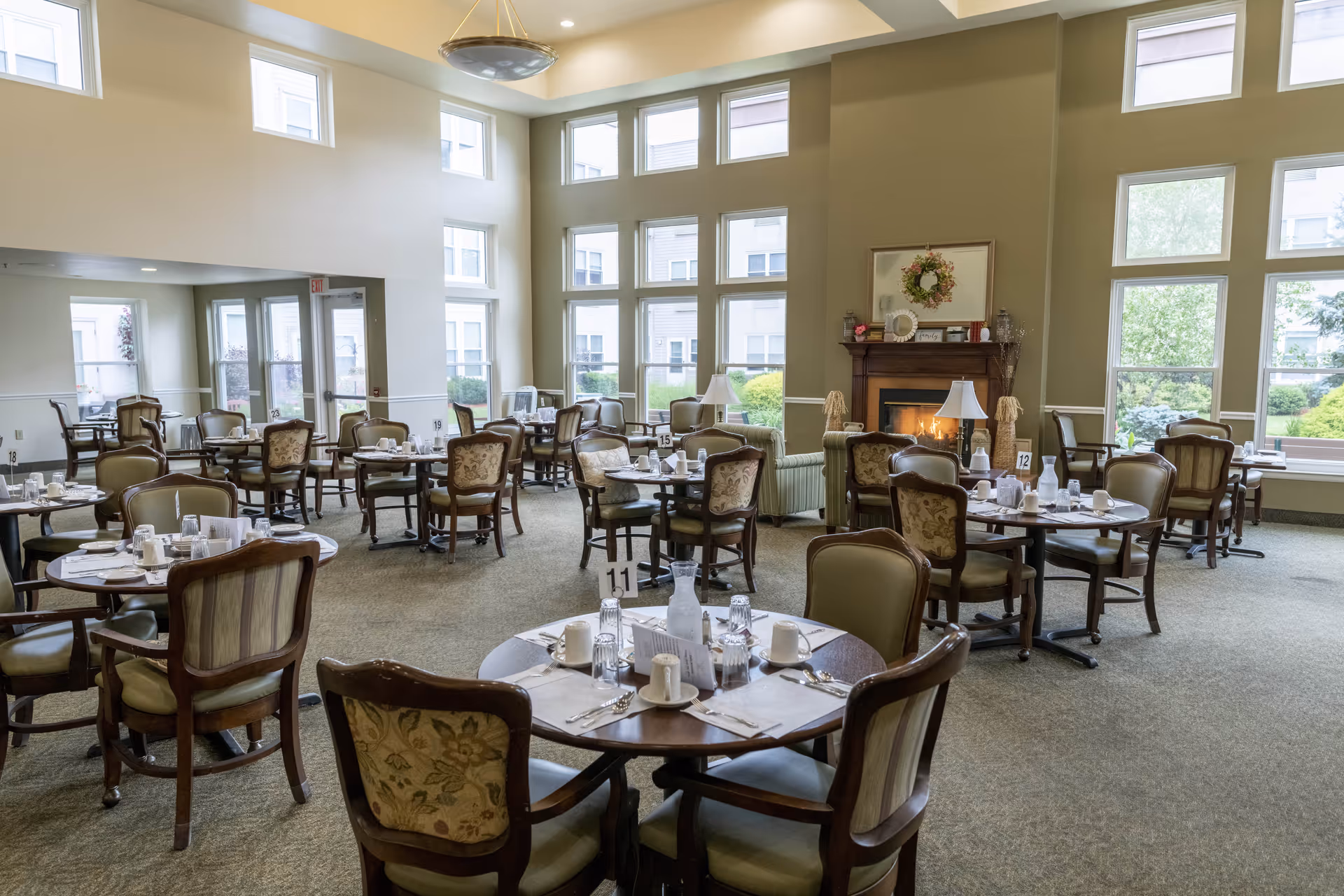 A spacious dining room with multiple round tables set with white cups, glasses, and silverware. The room features large windows letting in natural light, a fireplace with a wreath above it, and comfortable upholstered chairs around each table.