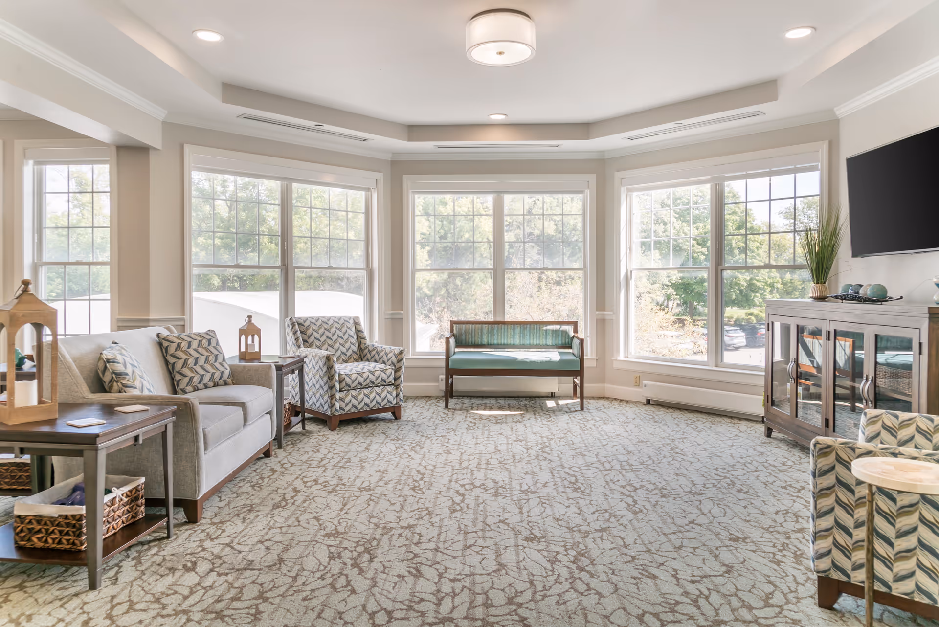 Bright furnished sitting room with large bay windows, sofas, patterned armchairs, a bench, and a TV cabinet.
