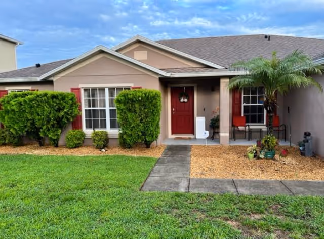 Front exterior of a single-story residence with a red door, windows with red shutters, porch chairs, shrubs, and a small palm tree.