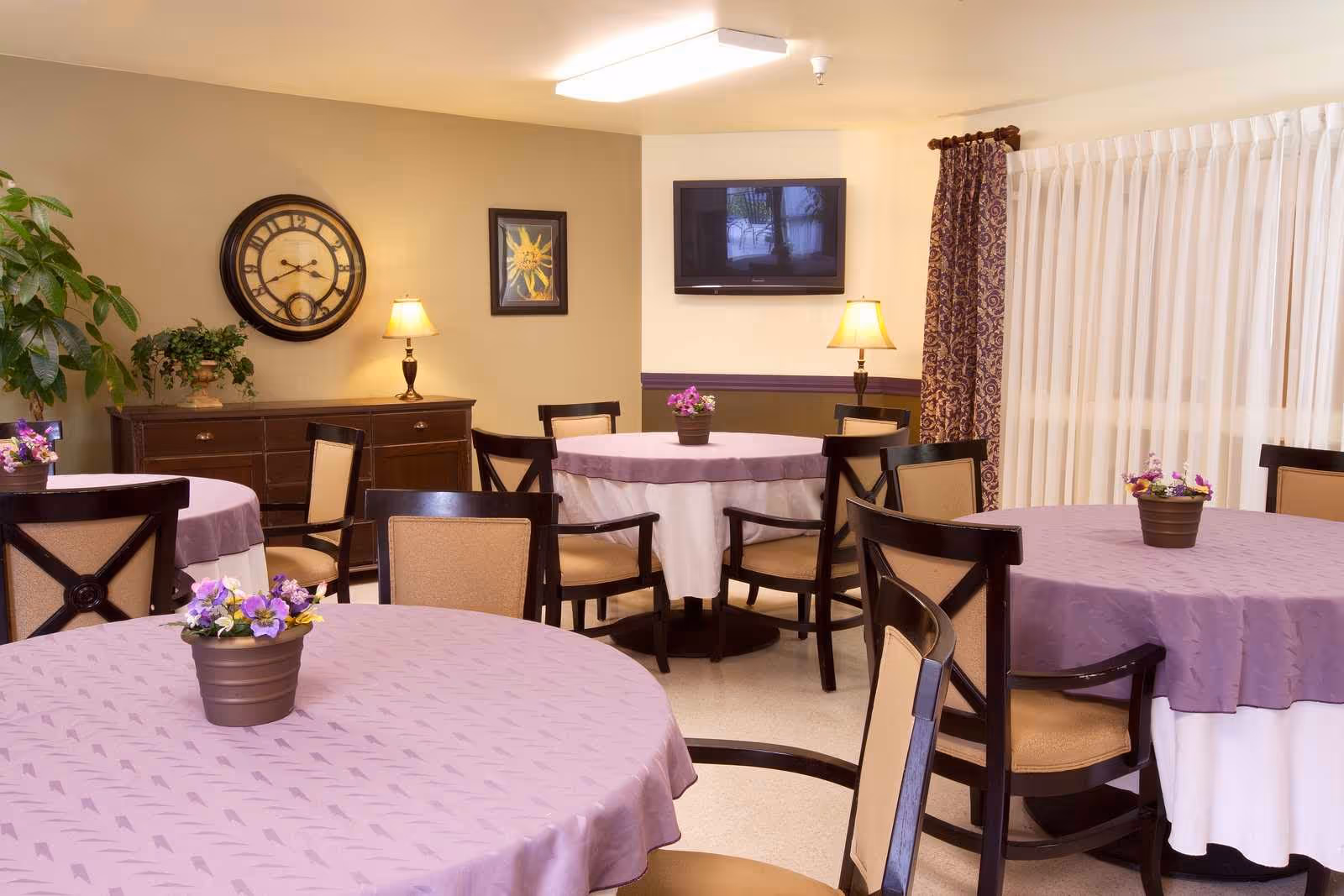 A dining room with round tables covered in lavender tablecloths, each with a small flower pot centerpiece. The room has several wooden chairs with beige cushions, a sideboard with a large wall clock and a framed picture above it, two table lamps, a flat-screen TV mounted on the wall, and large windows with sheer white curtains and patterned drapes.