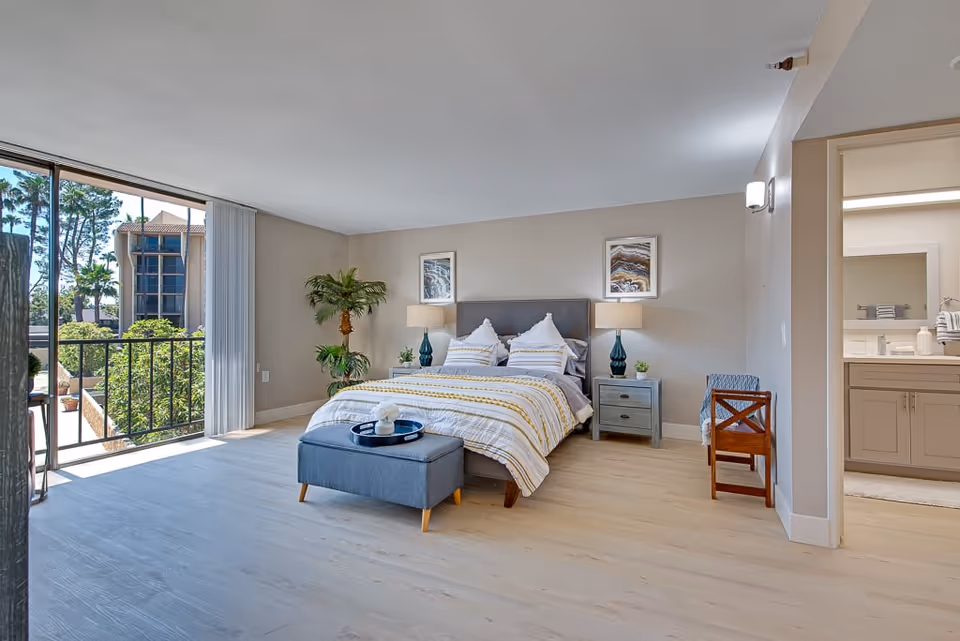 Spacious bedroom with a made bed, ottoman, nightstands, sliding glass doors to a balcony and an adjoining bathroom visible.