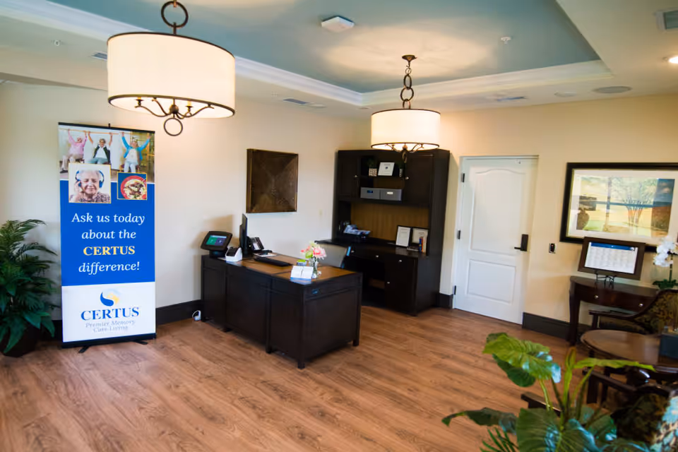 Reception area of CERTUS Premier Memory Care Living with a dark wooden desk, computer, and office chair. A blue and white promotional banner stands to the left, and there are two large hanging light fixtures on the ceiling. The room has wood flooring, a white door, framed artwork on the wall, and some green plants.