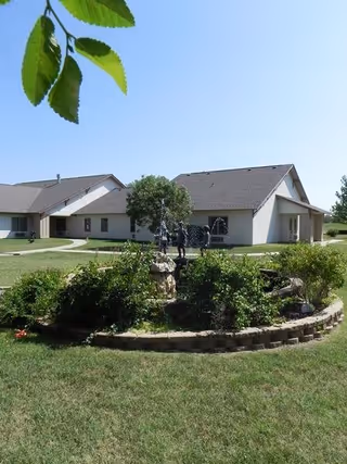 A landscaped garden area with green bushes and a small tree in the center, surrounded by a circular stone border. In the background, there is a single-story building with beige walls and a gray roof under a clear blue sky. A few leaves hang down from the top left corner of the image.