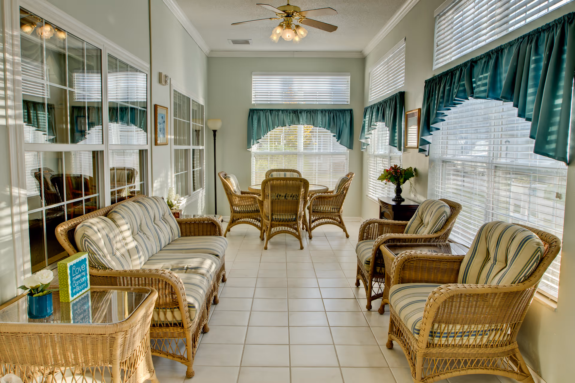 A bright sunroom with wicker furniture including a sofa, two armchairs, a glass-top side table, and a round dining table with four chairs. The room has large windows with white blinds and teal valances, a ceiling fan with lights, and tiled flooring. There is a small decorative sign on the side table that reads 'Love makes my garden bloom' and a vase with flowers on a side cabinet.