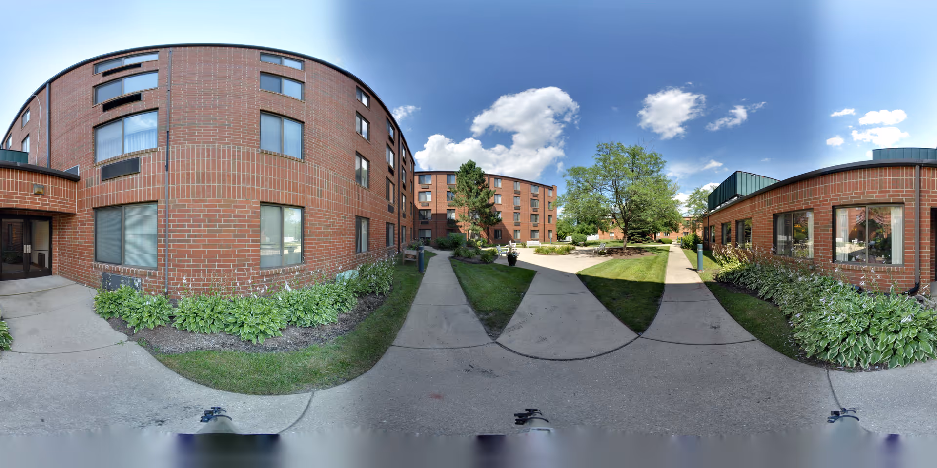 Outdoor courtyard area of a senior living facility with brick buildings on both sides, concrete walkways, green grass, and trees under a partly cloudy blue sky.