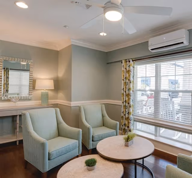 A cozy sitting area in a senior living facility with two light green armchairs, two round marble-top coffee tables with small potted plants, a large window with blinds and floral curtains, a ceiling fan with light, a wall-mounted air conditioning unit, a console table with a lamp, and a decorative mirror on the wall.