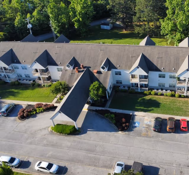Aerial view of a two-story senior living building with a covered entrance/porte-cochere, balconies, parking lot, and surrounding trees.