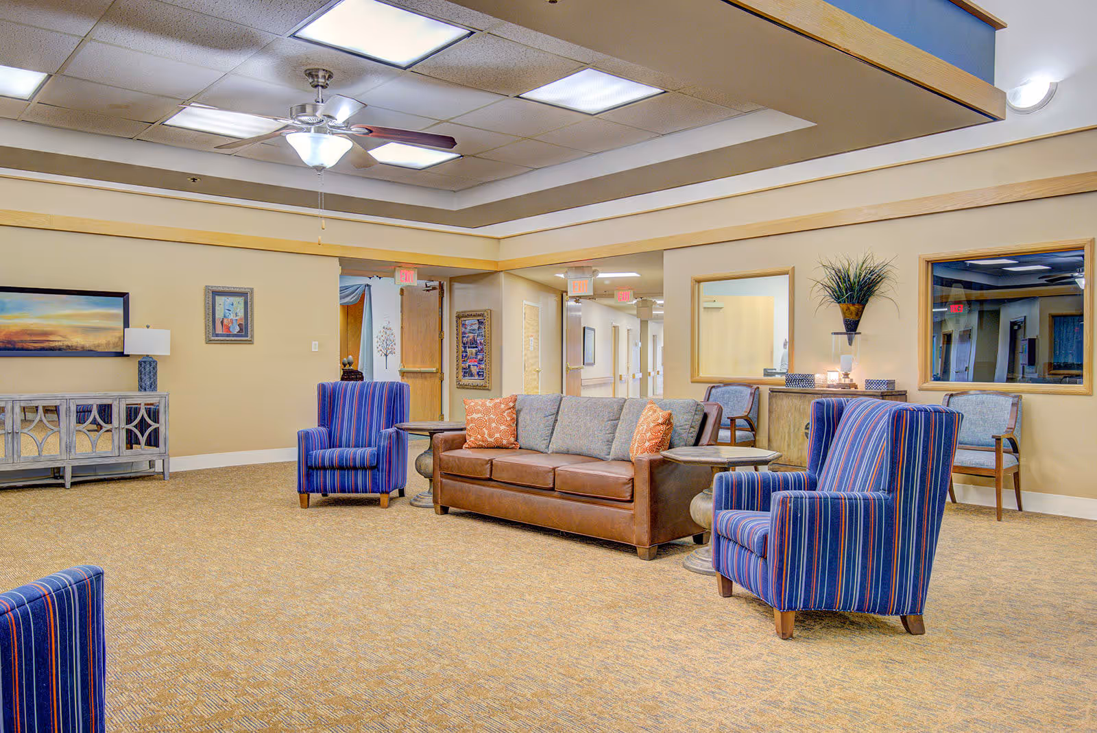 A spacious assisted living facility common area with a brown leather sofa, two blue striped armchairs, side tables, and beige carpet. The walls are light beige with framed artwork and a decorative cabinet. Ceiling lights and a ceiling fan illuminate the room. There are windows and doorways leading to a hallway with exit signs visible.