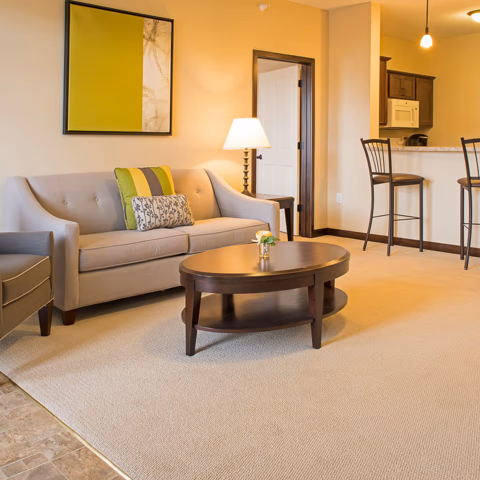 A cozy living room with a beige sofa adorned with two decorative pillows, a dark wooden oval coffee table with a small flower vase, a side table with a lamp, and two high chairs at a kitchen counter. The walls are painted light yellow, and there is a framed abstract artwork hanging above the sofa.