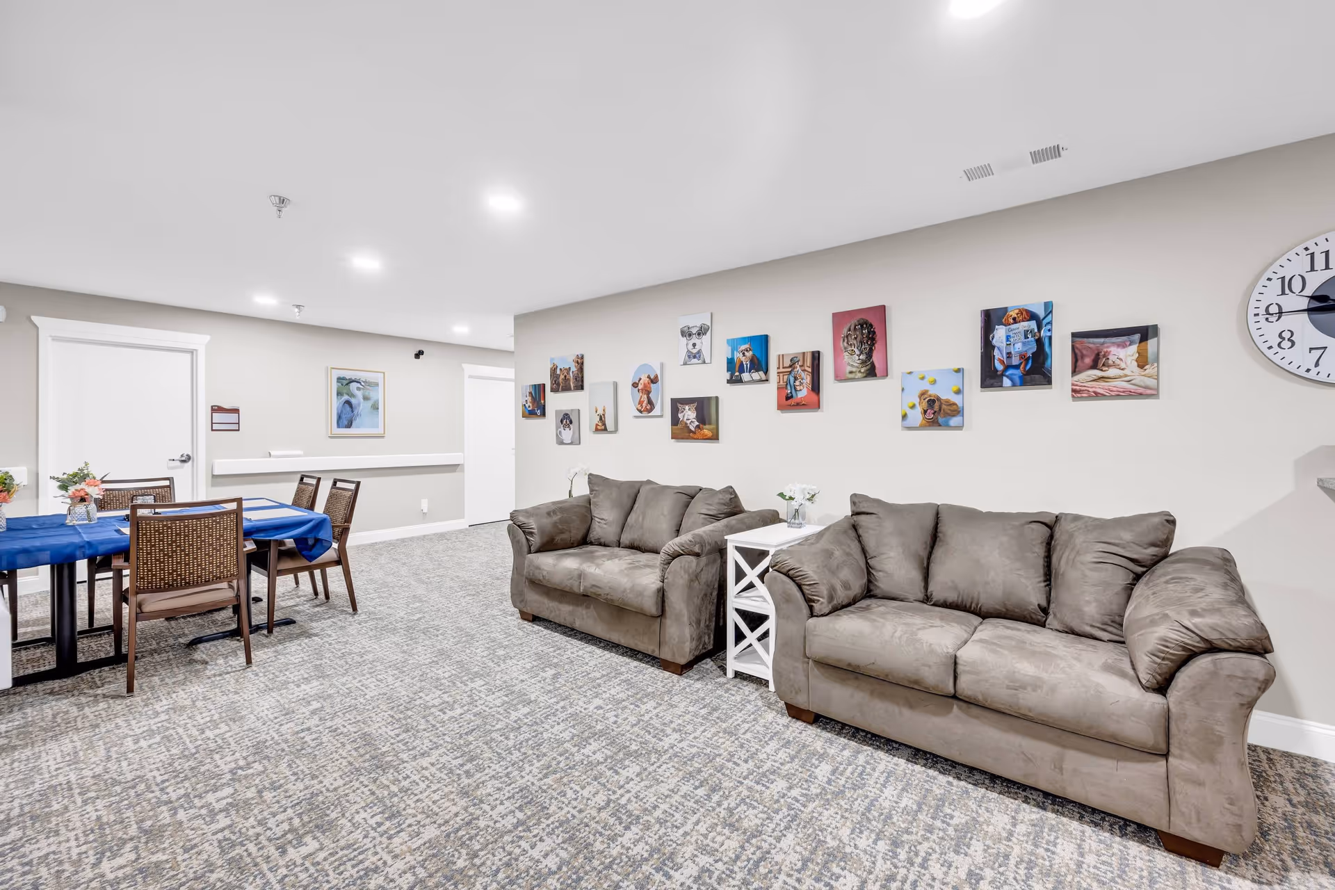 A cozy senior living common area with two gray sofas separated by a white side table with a flower vase. The wall behind the sofas is decorated with colorful animal paintings. To the left, there is a dining table covered with a blue tablecloth and surrounded by chairs. The room has light-colored walls, a patterned carpet, and a large clock on the wall.