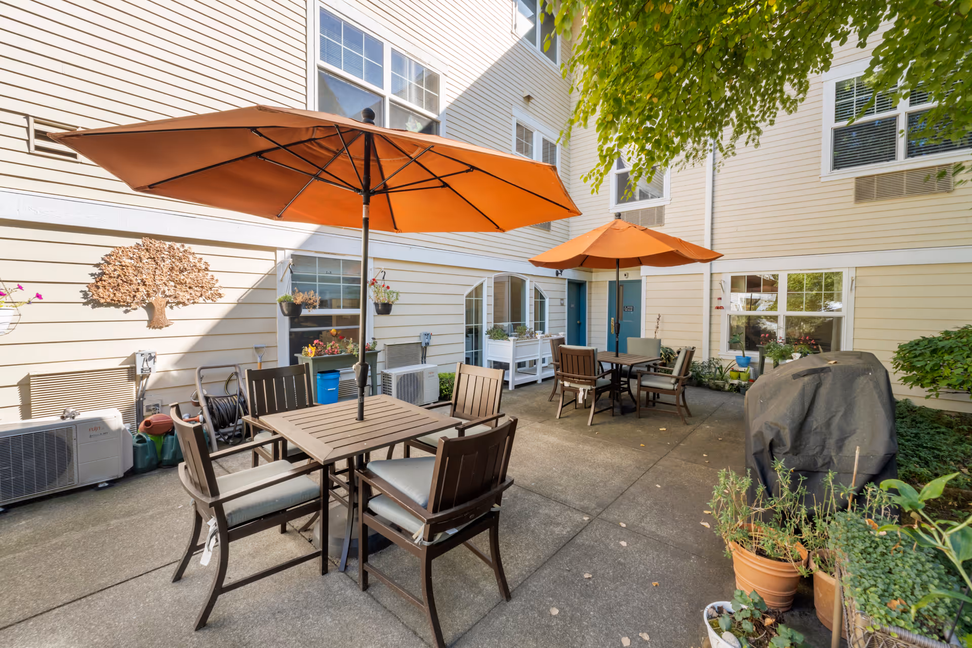 Outdoor patio area with two tables, each shaded by large orange umbrellas. Each table is surrounded by four cushioned chairs. The patio is adjacent to a light-colored building with multiple windows and doors. There are potted plants and a covered grill on the right side, with some garden tools and air conditioning units along the building wall.