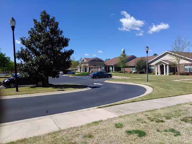 Front entrance of a senior living facility with a circular driveway, lawns, lampposts and parked cars under a blue sky.