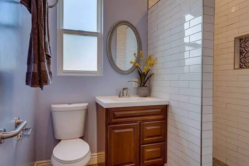 A small bathroom with a white toilet, a wooden vanity with a white countertop and a faucet, an oval mirror above the sink, a window with frosted glass, a towel hanging on the wall, and a shower area with white subway tiles and a decorative tile inset.