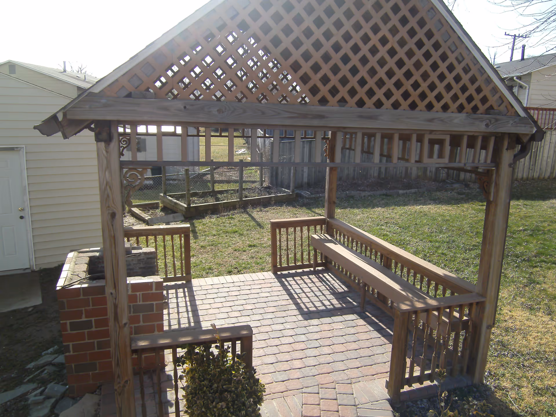 A wooden gazebo with lattice roof and built-in benches on a brick patio in a backyard. There is a small brick structure on the left side, possibly a grill or fireplace, and a grassy yard with a fence and neighboring houses in the background.