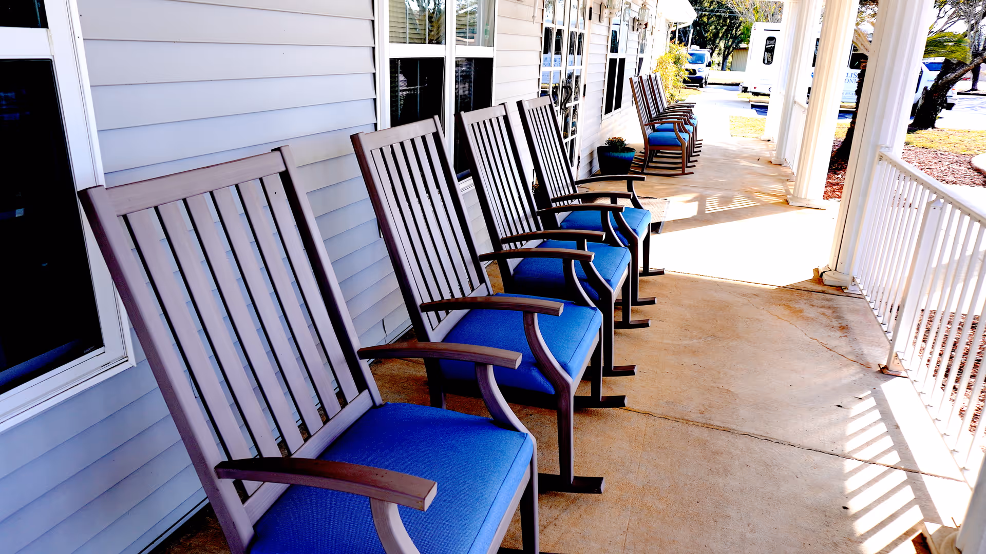 A row of wooden rocking chairs with blue cushions lined up on a covered porch outside a building with light gray siding and white trim. The porch has a concrete floor and white railing, with sunlight casting shadows on the ground.