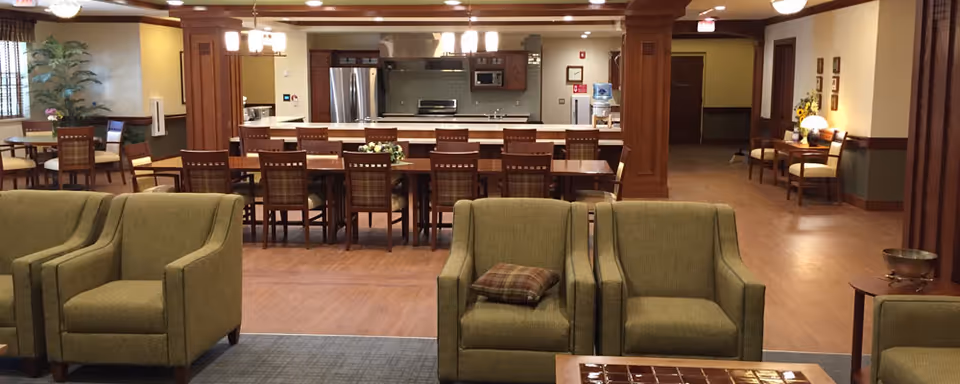 Open communal lounge and dining area with green armchairs in the foreground and a long dining table and kitchen in the background.