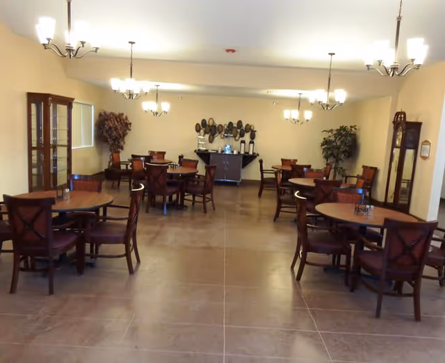 A spacious dining room with several round wooden tables and chairs arranged neatly. The room is well-lit with multiple chandeliers hanging from the ceiling. There are decorative plants and a sideboard with coffee urns and cups against the far wall.