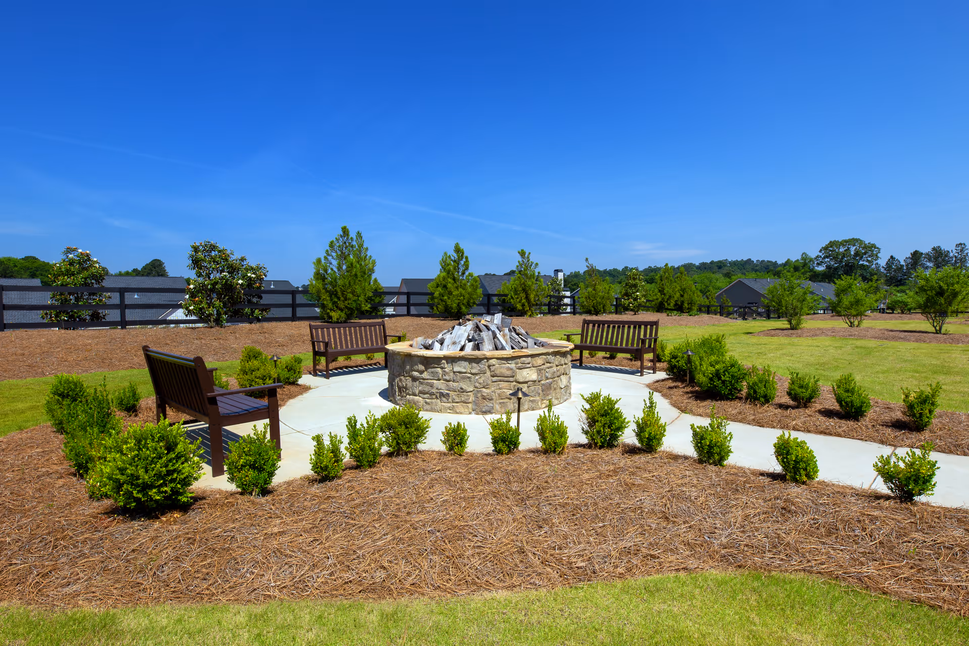 Outdoor seating area with four wooden benches arranged around a circular stone fire pit, surrounded by small green bushes and landscaped mulch, under a clear blue sky.