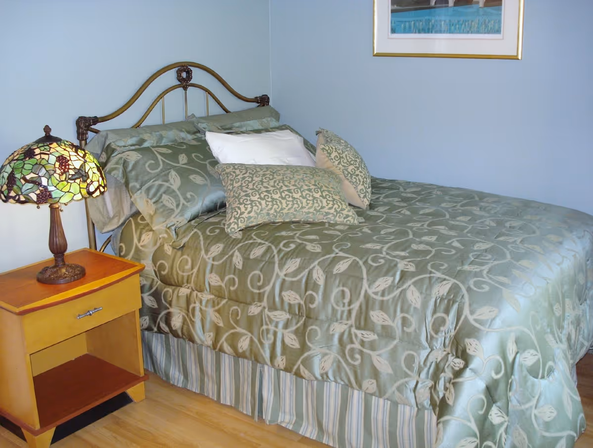 A bedroom with a neatly made bed featuring a green patterned bedspread and multiple pillows. Next to the bed is a wooden nightstand with a decorative stained glass lamp. A framed picture hangs on the light blue wall above the bed.