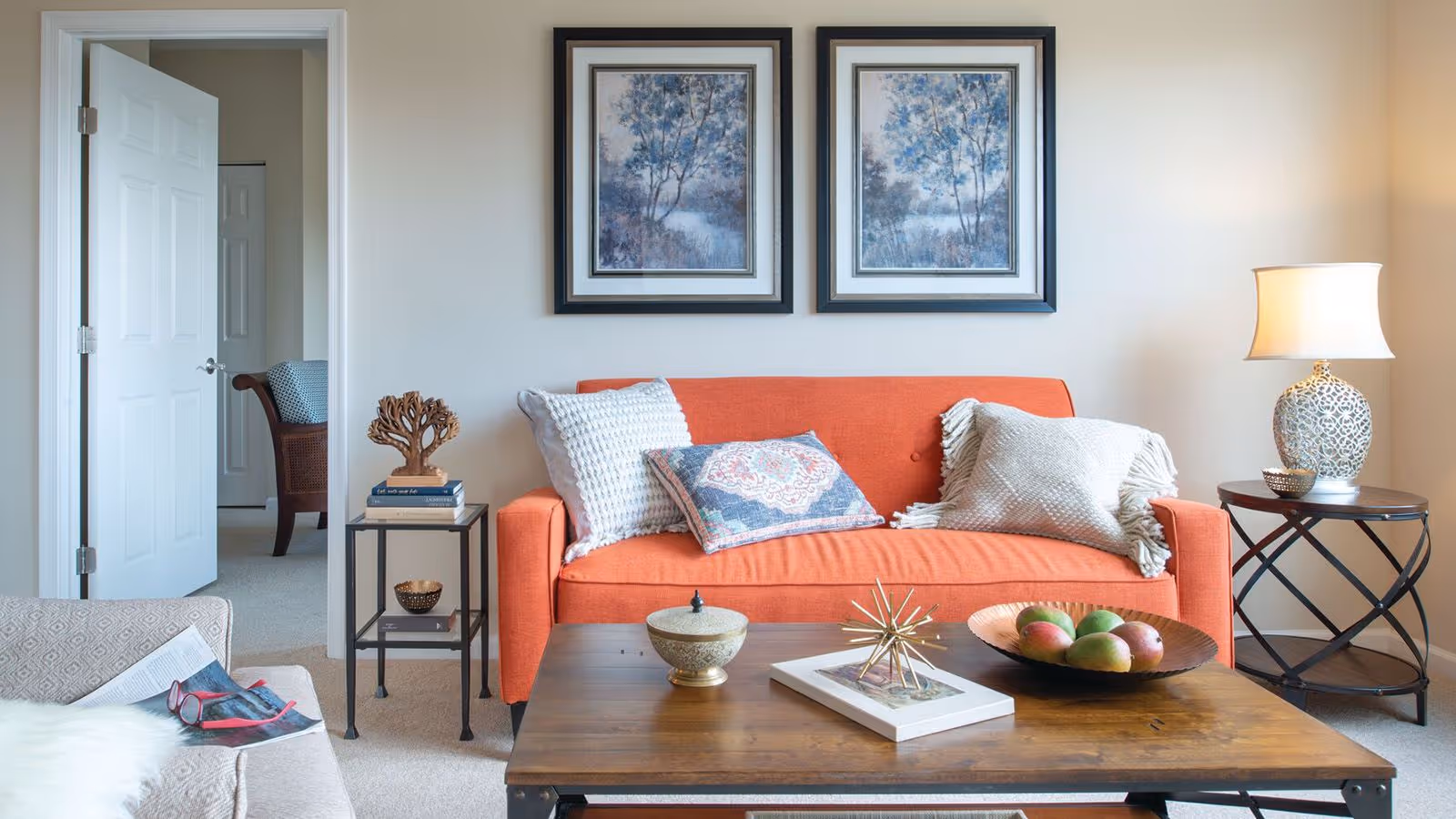 Bright living room with an orange sofa, patterned pillows, framed artwork, a wooden coffee table with decor and a side lamp.
