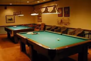 Interior room with two green felt pool tables, a brown couch along the wall, framed pictures on the walls, and overhead lighting fixtures above the pool tables.