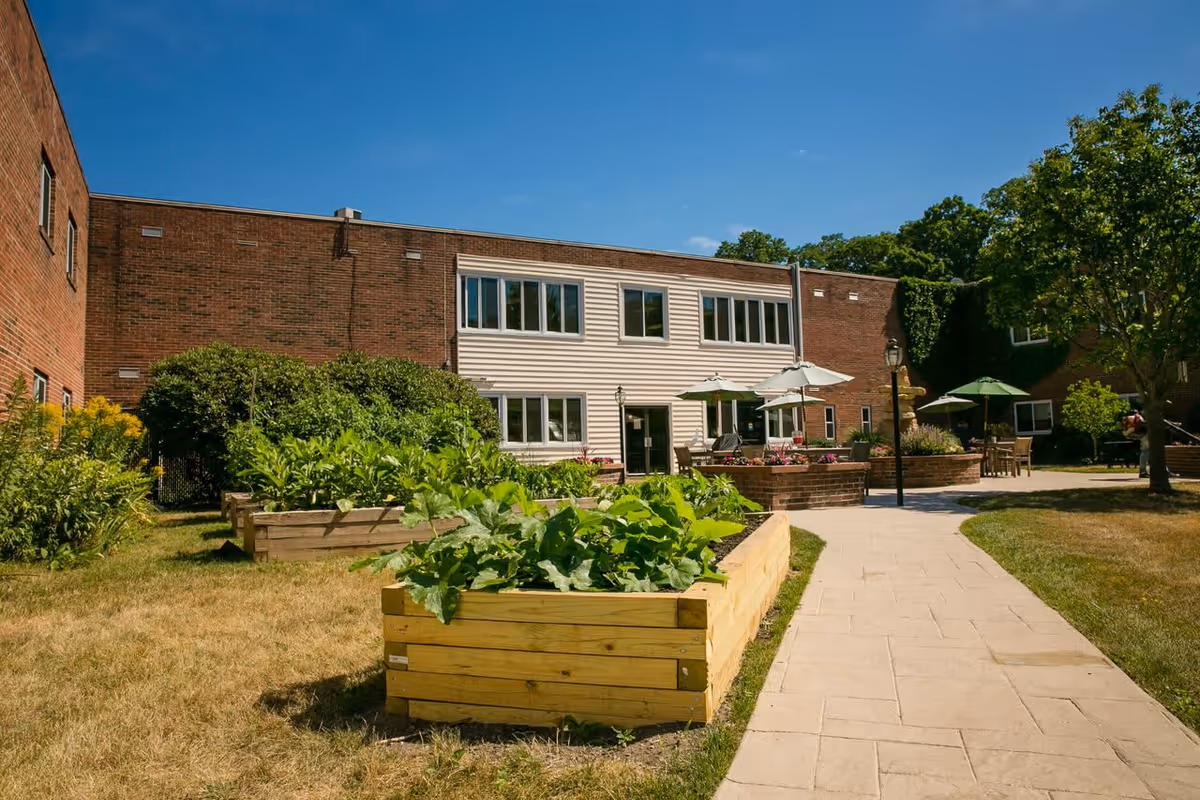 Outdoor courtyard area of a senior living facility with raised garden beds filled with green plants, a paved walkway, patio tables with umbrellas, and a brick building in the background under a clear blue sky.