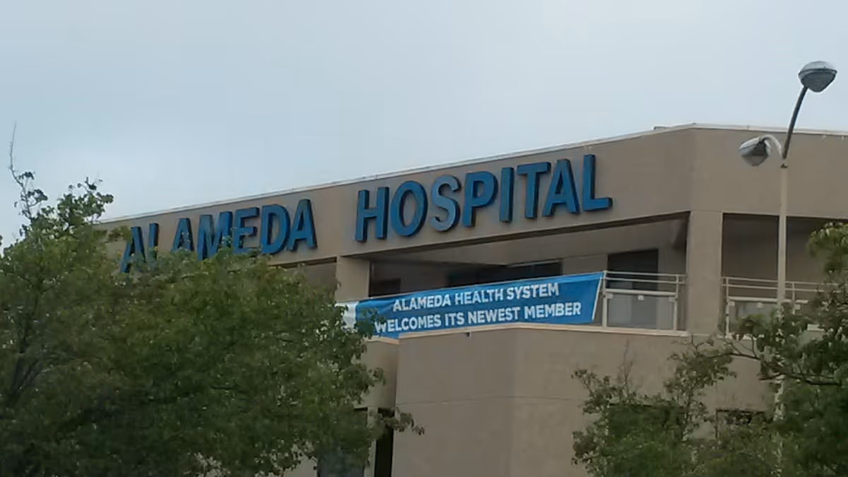 Exterior view of a beige building with large blue letters spelling 'ALAMEDA HOSPITAL' near the roofline. There is a blue banner hanging below that reads 'ALAMEDA HEALTH SYSTEM WELCOMES ITS NEWEST MEMBER'. Trees partially obscure the lower part of the building, and two street lamps are visible on the right side.