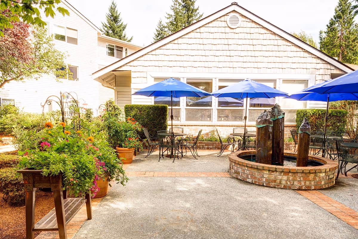 Outdoor patio area at The Gardens at Marysville featuring several metal tables and chairs with blue umbrellas, a circular brick water fountain with three vertical sculptures, and lush greenery including potted plants and flowers along the side.
