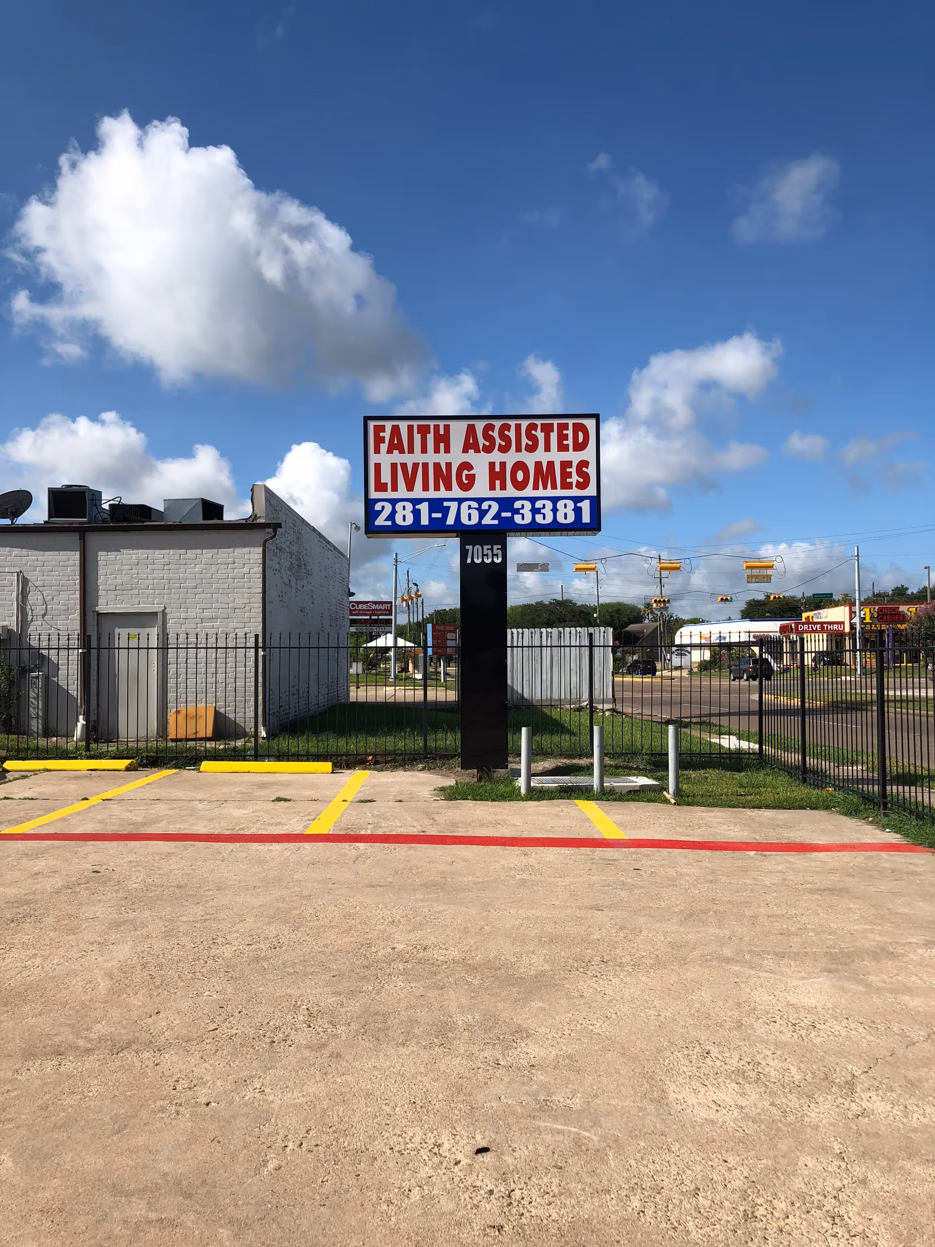 Outdoor view of a parking lot with a sign that reads 'Faith Assisted Living Homes' along with a phone number 281-762-3381. The sign is mounted on a black pole with the number 7055 on it. There is a fenced area and a white building in the background under a blue sky with scattered clouds.
