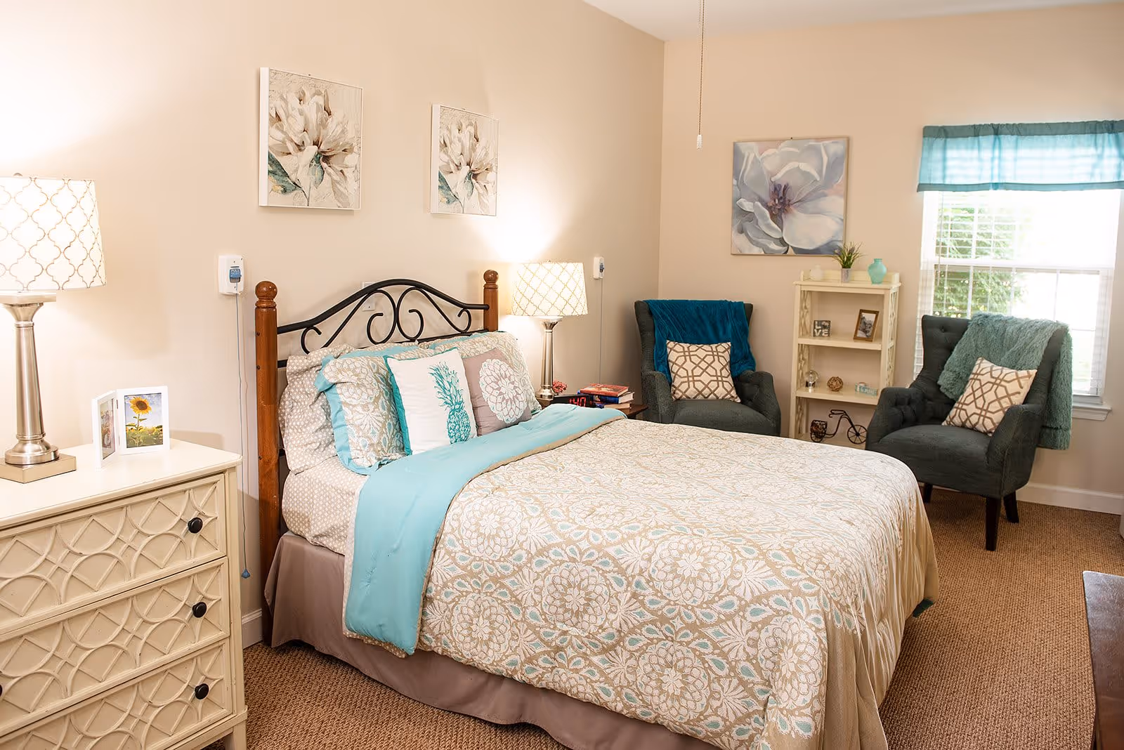 A cozy bedroom with a bed featuring a decorative headboard, beige and turquoise bedding, two bedside lamps, a white dresser with a small framed picture, two armchairs with patterned pillows, a small white bookshelf, and a window with turquoise curtains letting in natural light.