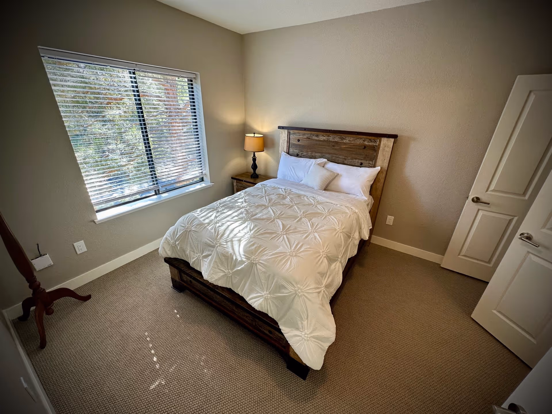 A cozy bedroom with a wooden bed frame and a white comforter. There are two pillows on the bed and a small lamp on a nightstand next to the bed. A window with blinds lets in natural light, and the room has beige walls and carpeted flooring. A door is partially open on the right side of the room.