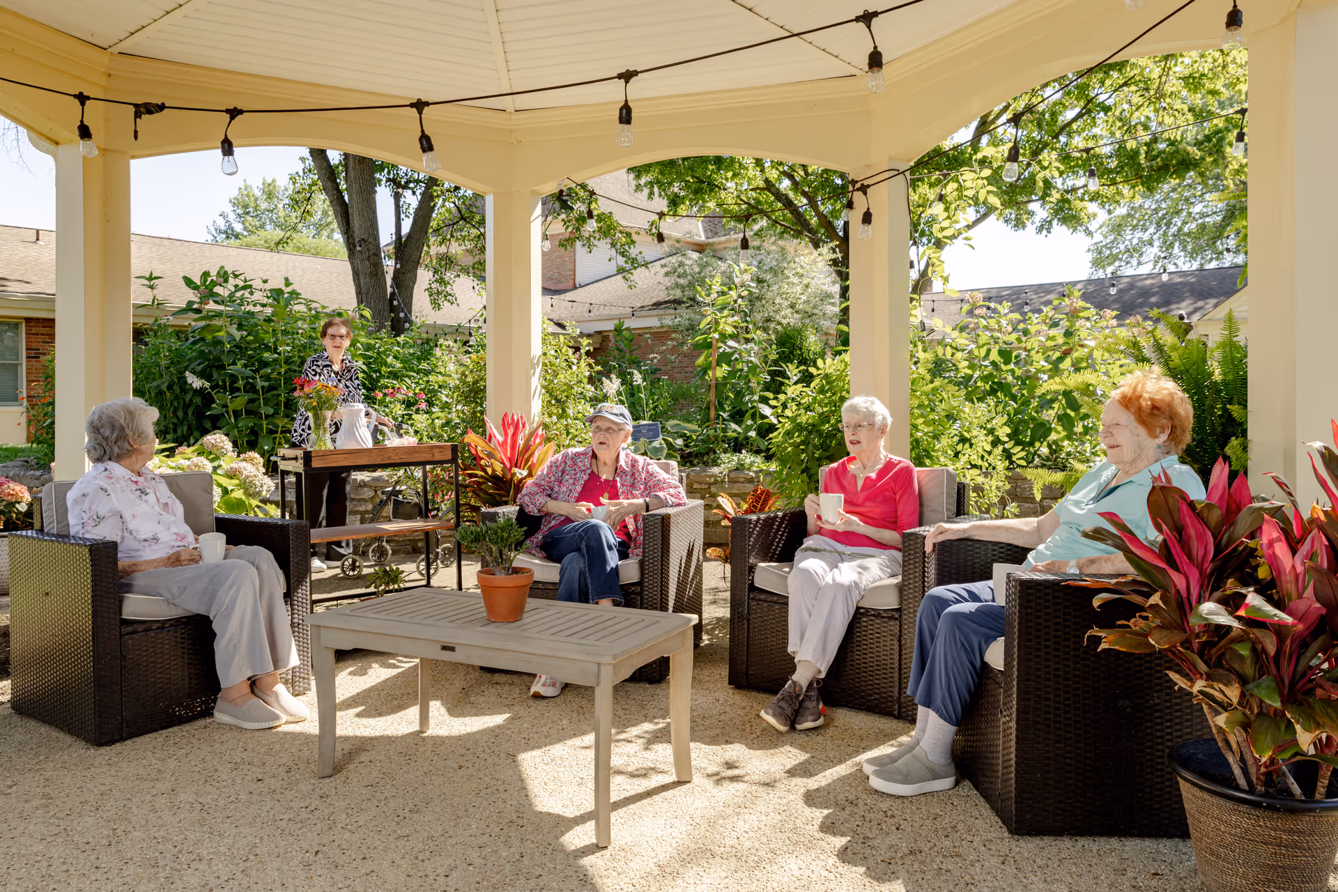 Four elderly women sitting on outdoor wicker chairs under a gazebo, enjoying drinks and conversation. A fifth woman stands behind a serving cart with flowers and a pitcher. The setting is a sunny garden area with lush green plants and trees surrounding the space.