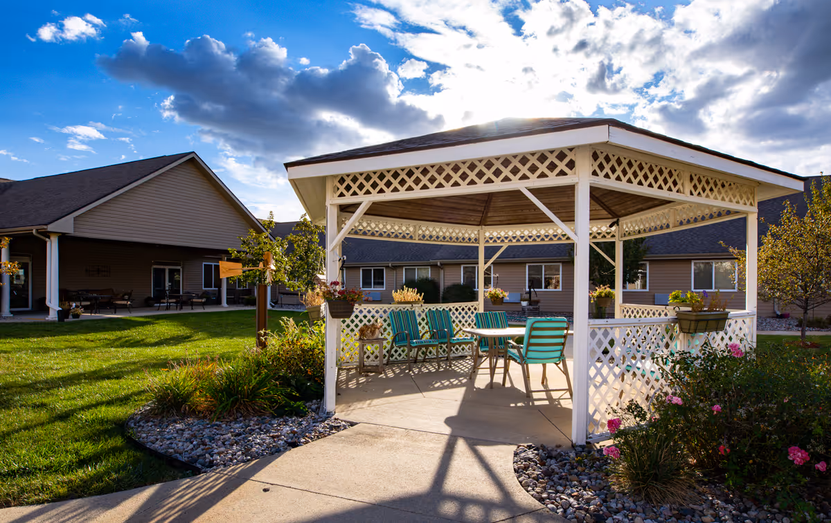 Outdoor gazebo with white lattice structure and green-striped chairs around a table, surrounded by a garden with flowers and shrubs, with senior living facility buildings in the background under a partly cloudy sky.