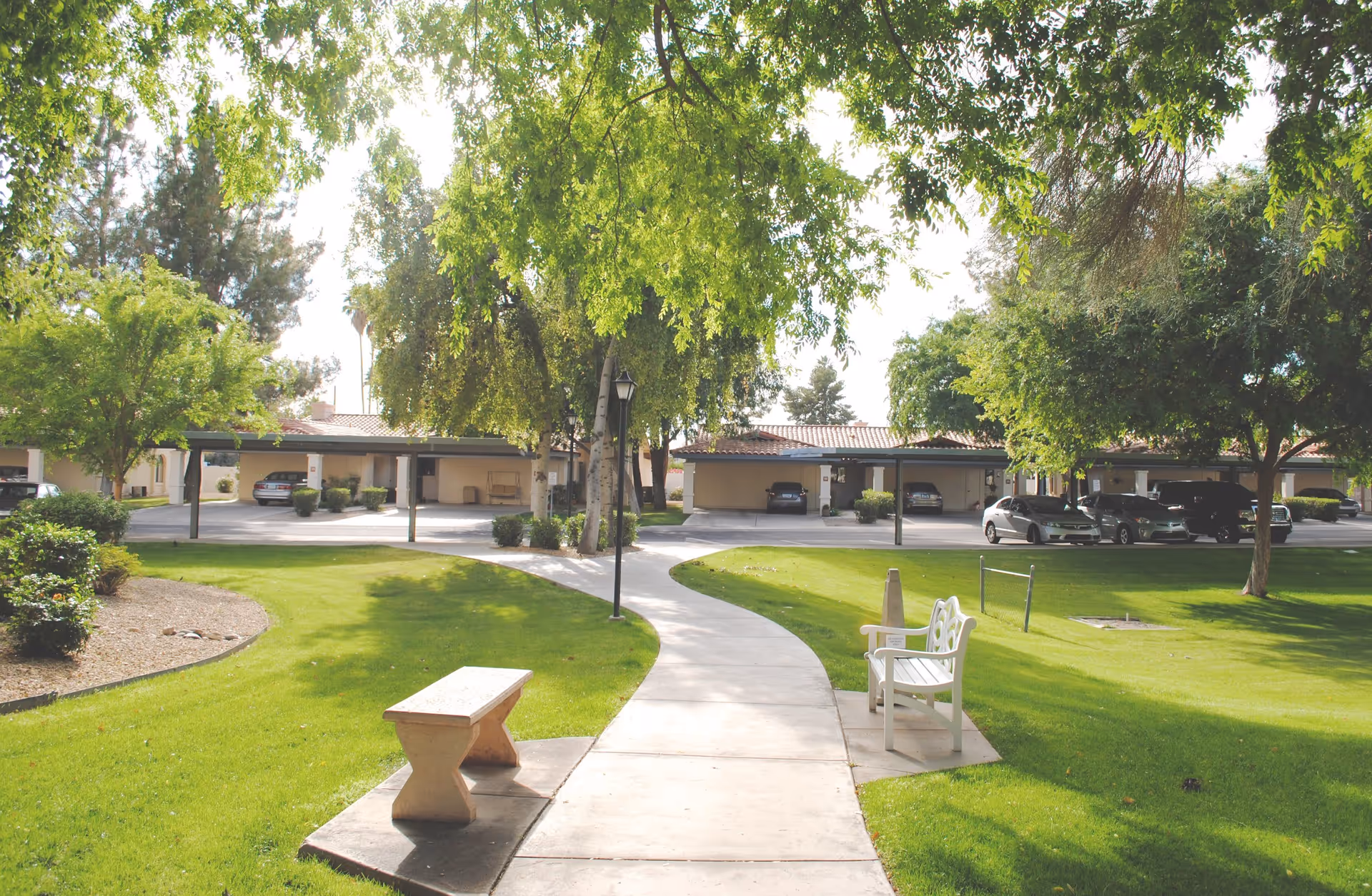 A sunny outdoor pathway in a senior living facility with green grass, trees providing shade, a stone bench on the left, and a white wooden bench on the right. In the background, there are covered parking spaces with several parked cars.