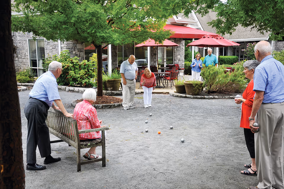 A group of elderly people playing bocce ball outdoors on a gravel court surrounded by trees and shrubs. Some are standing while one woman is sitting on a wooden bench. In the background, there is a building with stone walls, large windows, and red umbrellas shading patio tables and chairs.