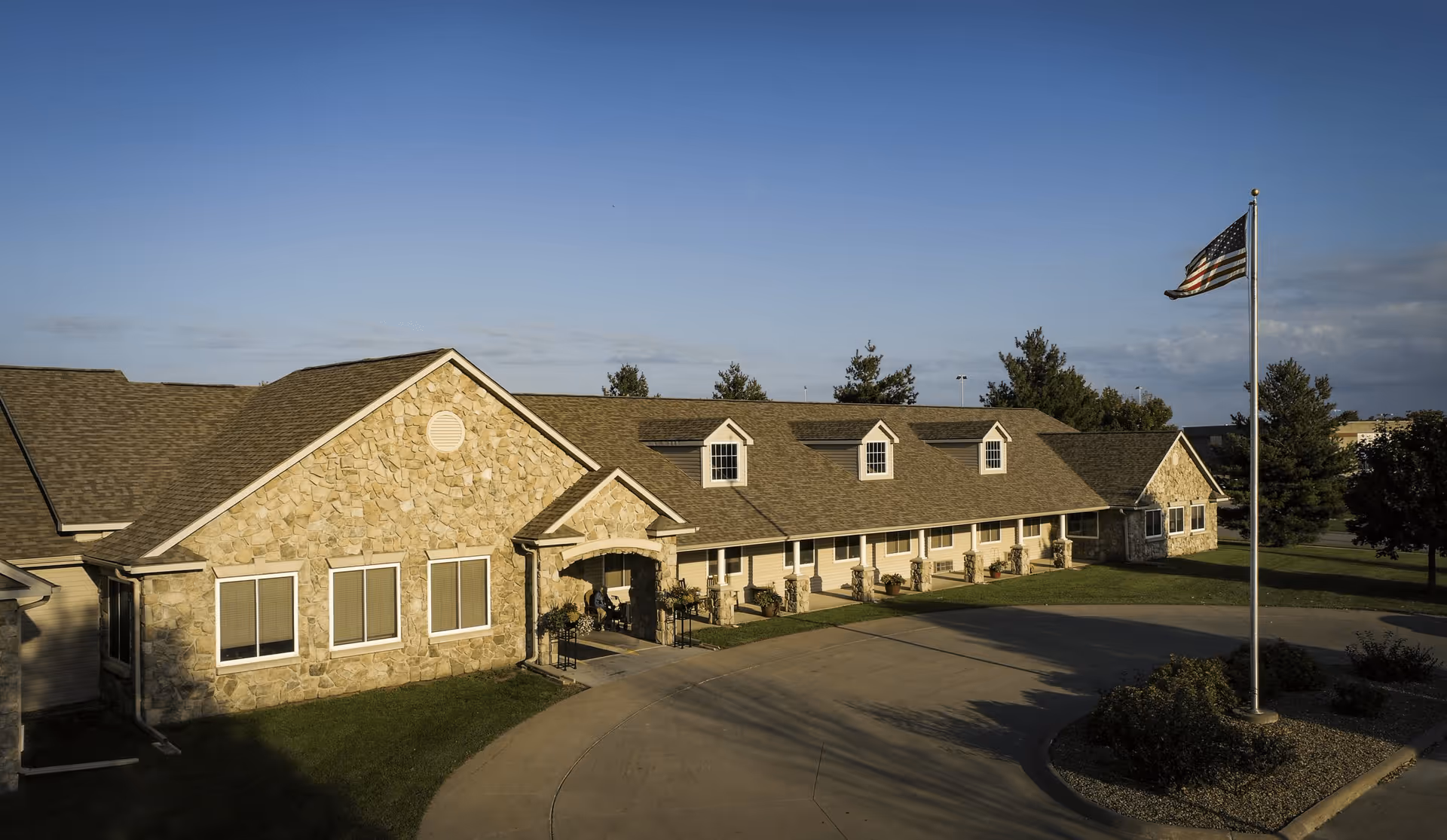 Exterior view of a single-story assisted living facility building with stone and siding facade, multiple windows, a covered entrance, and an American flag on a flagpole in front. The building is surrounded by grass, trees, and a curved driveway.