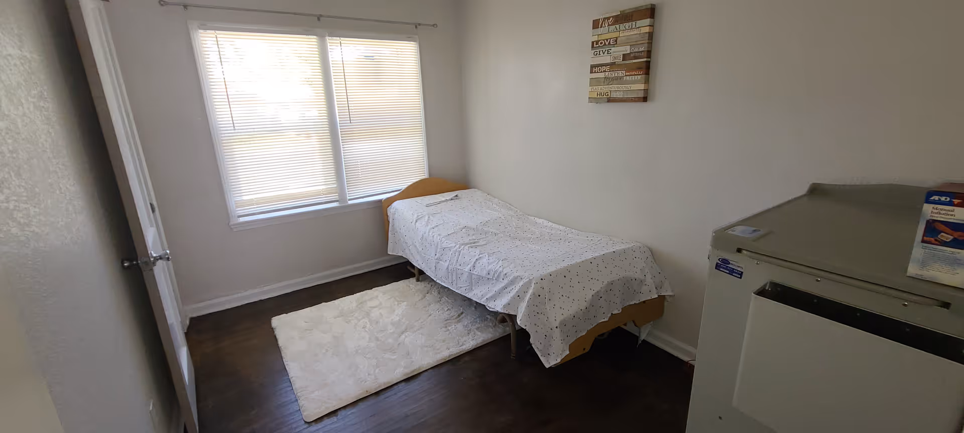 Small private bedroom with a single bed covered by a white sheet, a window with blinds, wall art, a rug on dark wood flooring, and a cabinet.
