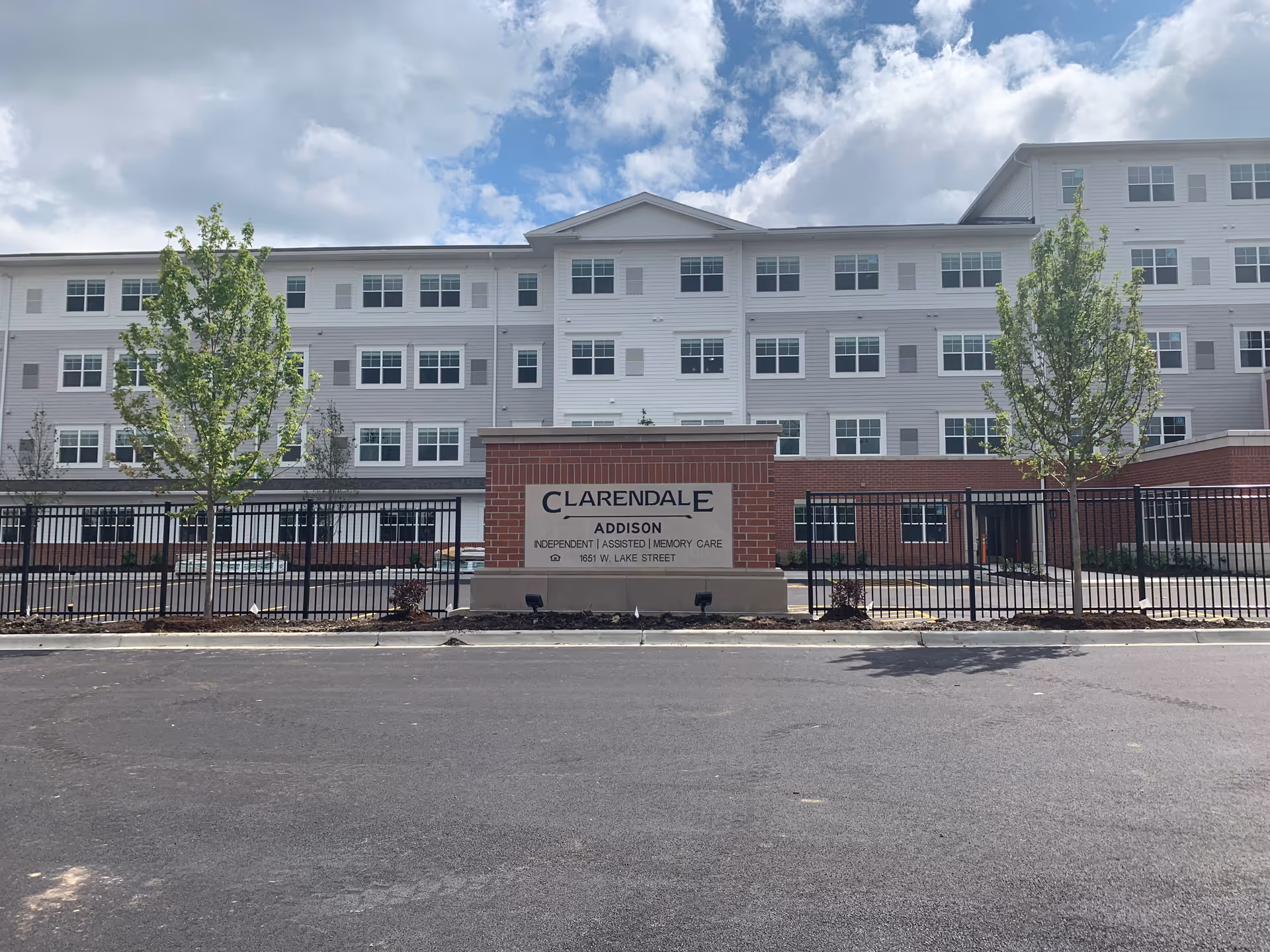 Front exterior view of Clarendale of Addison, a multi-story senior living facility with a brick and white facade, a sign in front displaying the facility name and services, and a black metal fence with trees on either side under a partly cloudy sky.