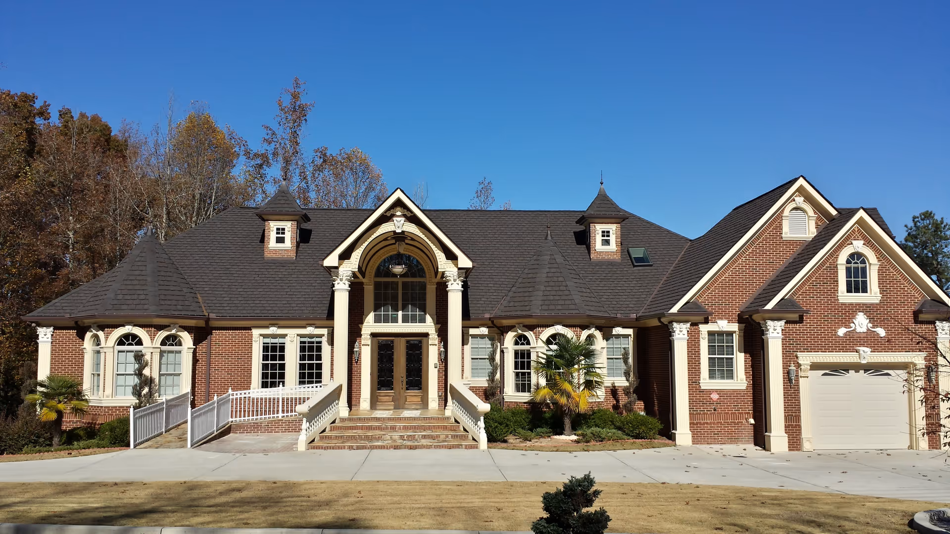 Front exterior view of a large, elegant brick building with a dark shingled roof, decorative white columns, arched windows, and a central entrance with steps and a wheelchair ramp. The building is surrounded by a well-maintained lawn and some trees under a clear blue sky.