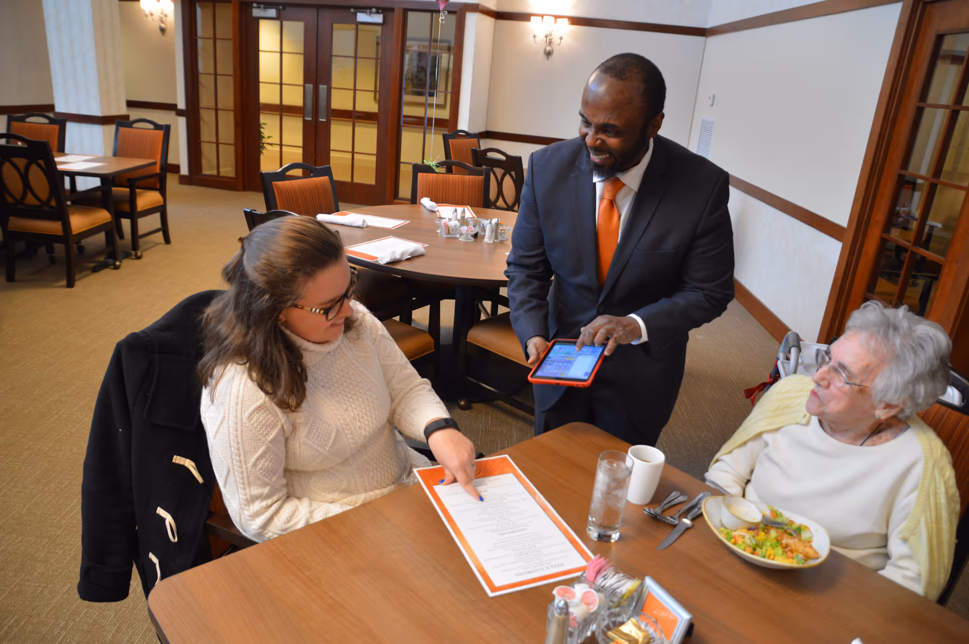 A man in a suit with an orange tie is standing and holding a tablet, interacting with two women seated at a dining table. One woman is pointing at a menu, and the other elderly woman has a plate of food in front of her. The setting appears to be a dining room with tables and chairs in the background.