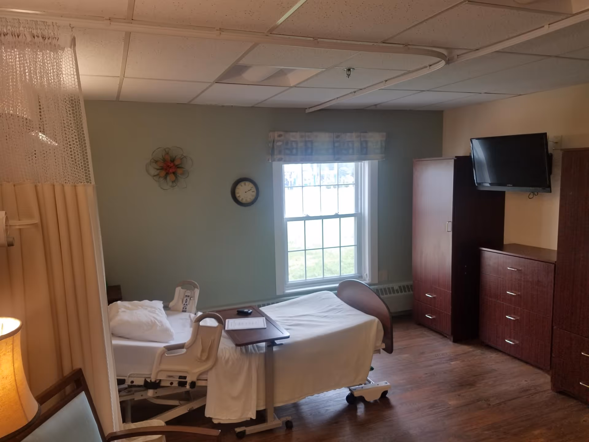 A senior living facility bedroom with a hospital-style bed, a small overbed table, a pillow, and a blanket. The room has a window with a patterned valance, a wall clock, a decorative wall piece, a mounted flat-screen TV, and wooden furniture including a dresser and a wardrobe. There is a privacy curtain partially visible on the left side and a wooden chair with a cushion in the foreground.