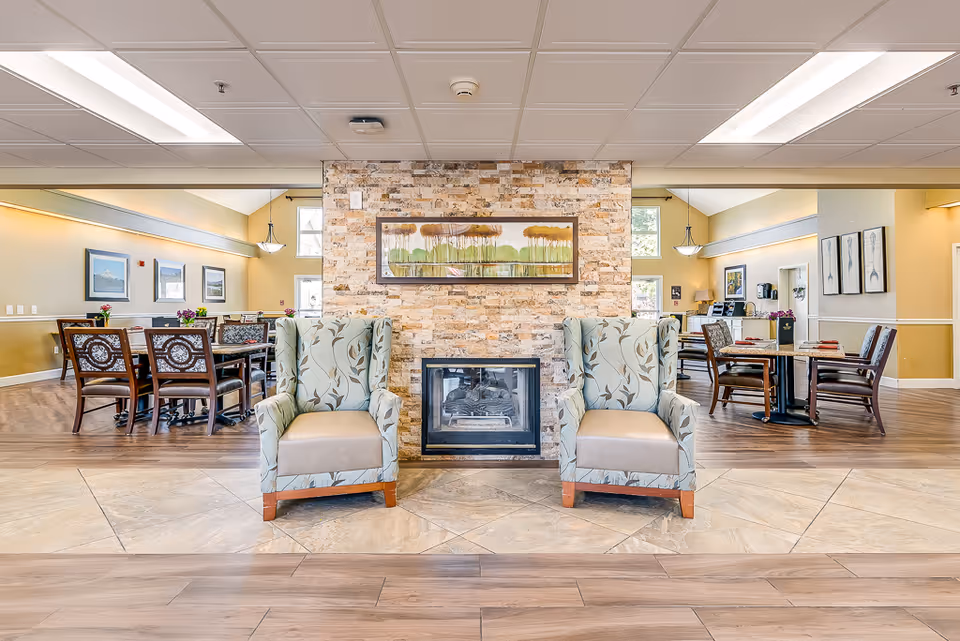 Bright senior living communal dining and lounge area with two upholstered armchairs flanking a central stone fireplace and dining tables in the background.