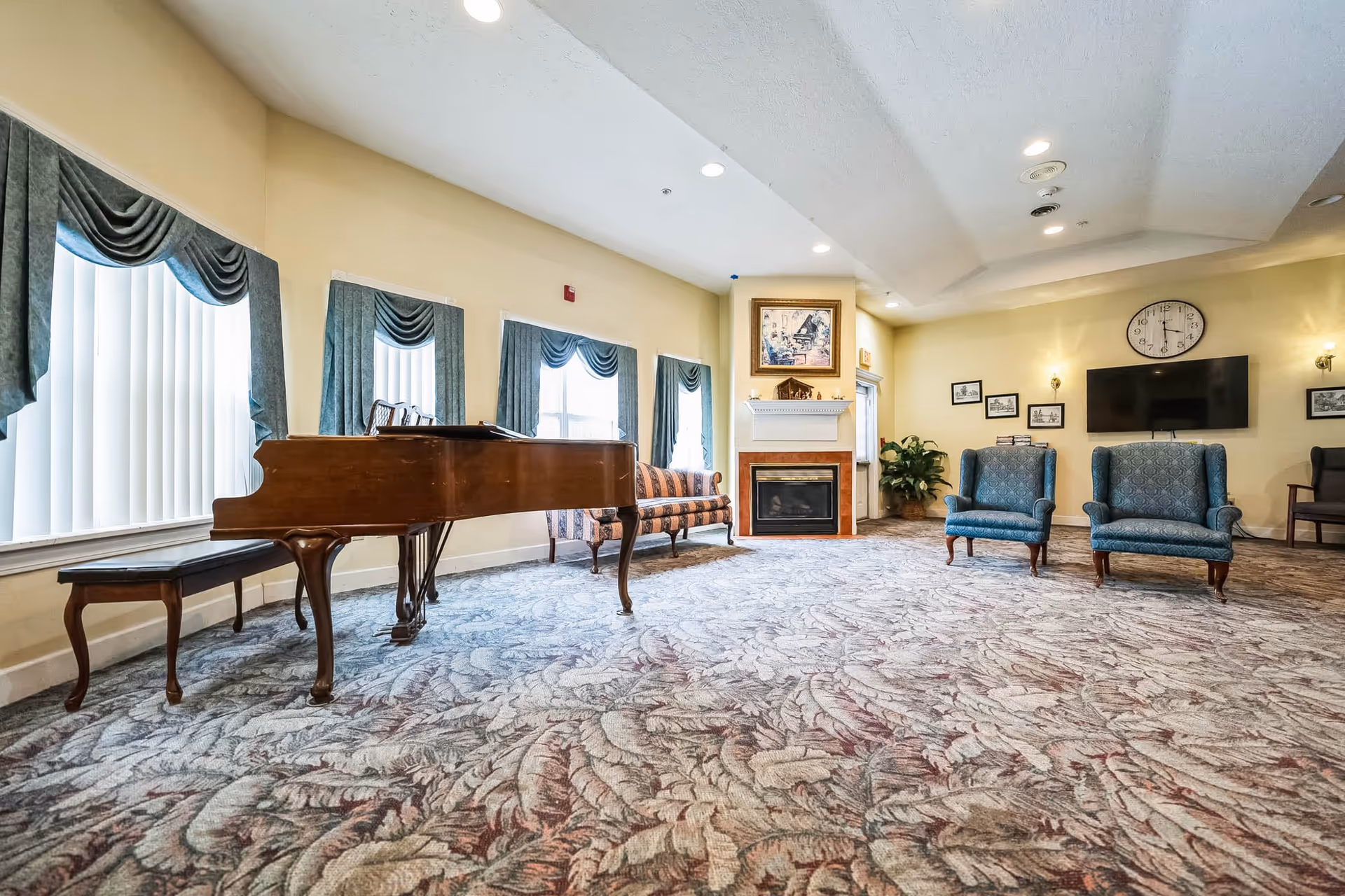A spacious living room with a patterned carpet, a grand piano with a matching bench, a striped sofa, two blue upholstered armchairs, a fireplace with a framed picture above it, a wall-mounted TV, a clock, and several framed pictures on the walls. The room has large windows with blue curtains and vertical blinds.