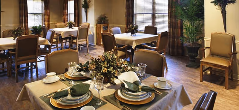 A dining room with multiple tables set with plates, napkins, glassware, chairs, and potted plants.