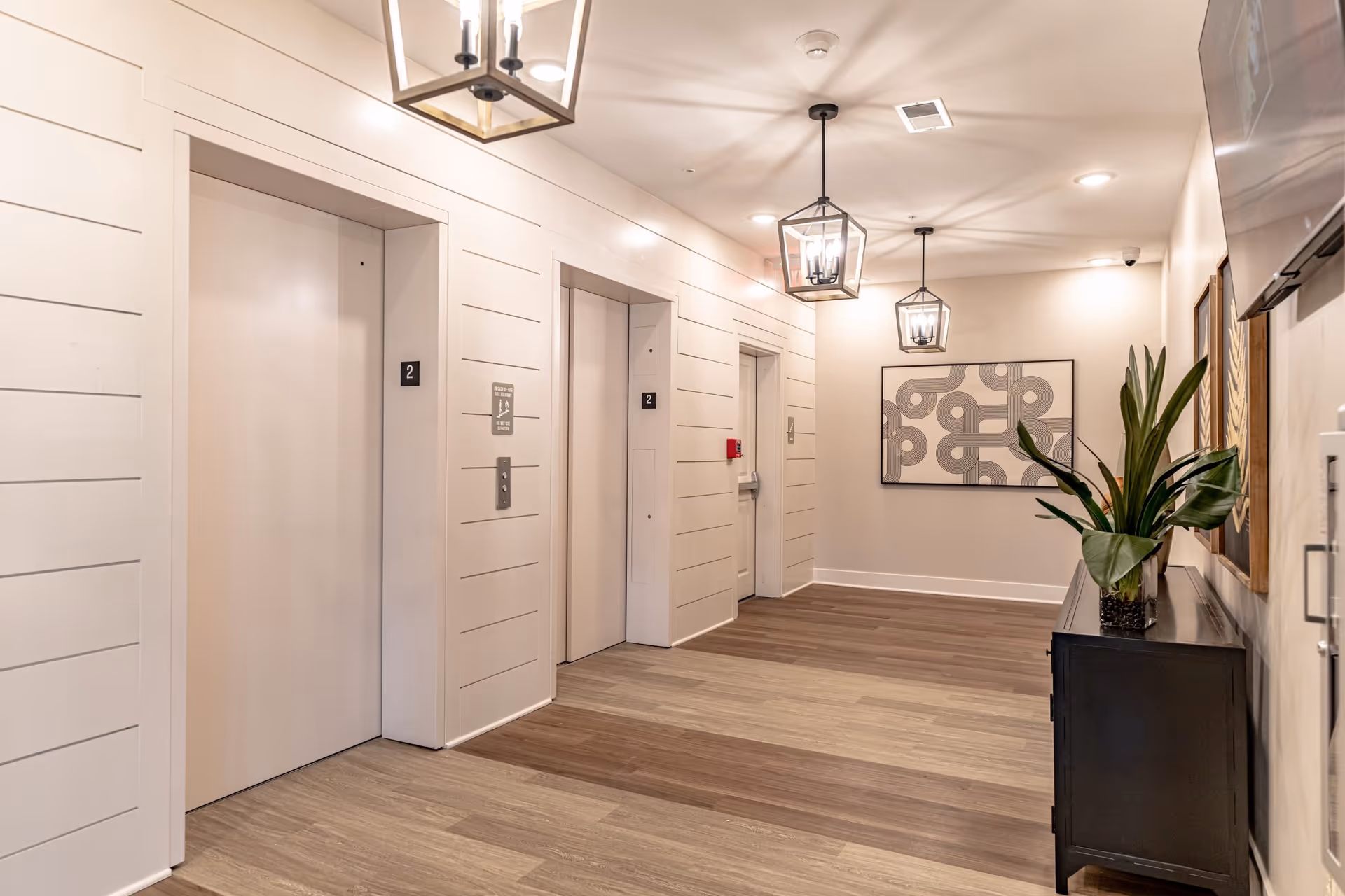 Well-lit interior hallway with two elevators, pendant lights, artwork, and a console table with a plant.