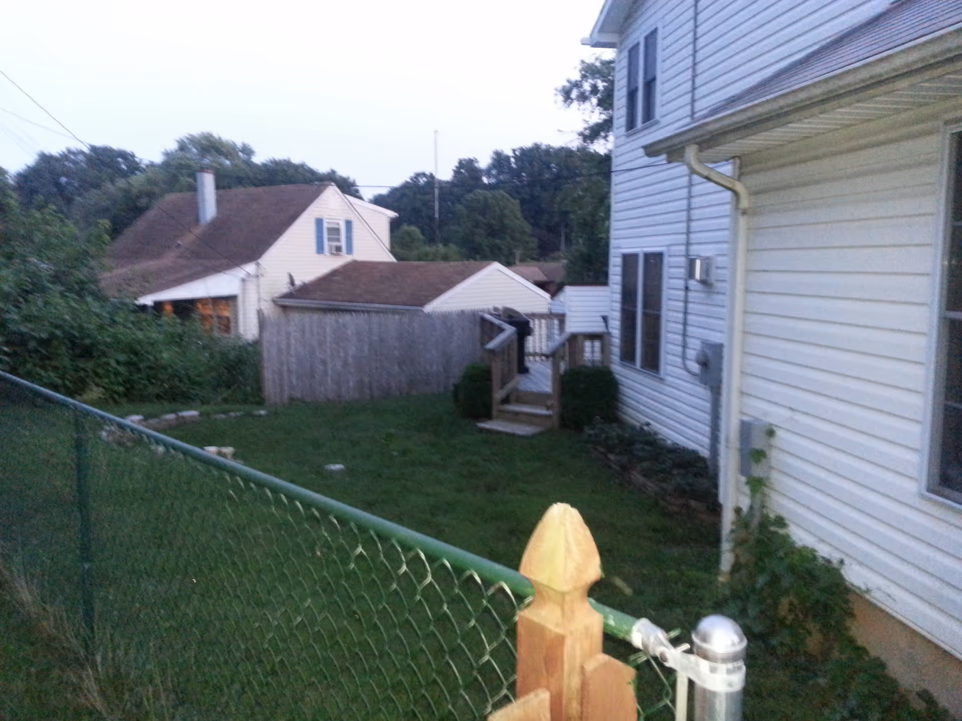 A backyard area with a green chain-link fence in the foreground, a grassy lawn, and a white house with siding on the right side. There is a small wooden staircase leading to a door on the side of the house. In the background, there are other houses and trees.