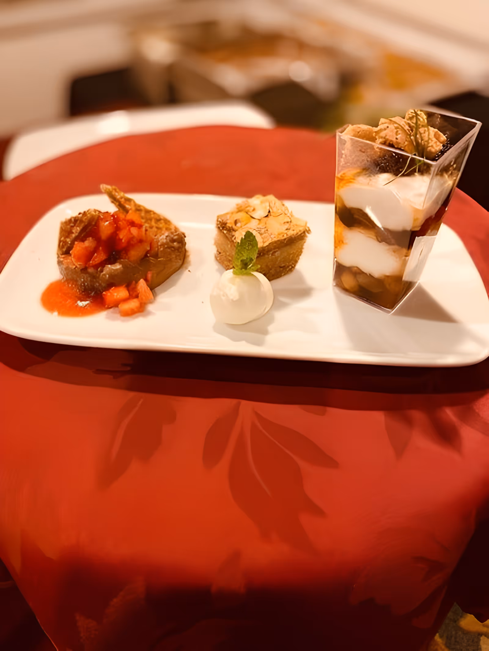 A white rectangular plate with three dessert items placed on a red tablecloth. On the left is a dessert topped with diced strawberries and a red sauce, in the middle is a square almond-topped pastry with a small dollop of cream garnished with a mint leaf, and on the right is a layered dessert in a clear plastic cup with cream, chocolate, and cookie pieces.