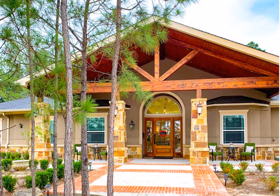 Front entrance of Village Green Memory Care Community Tomball featuring a covered porch with wooden beams, stone pillars, glass double doors, outdoor seating with tables and chairs, and surrounded by trees and landscaping.