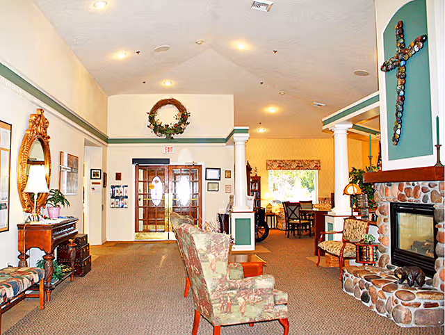 A cozy assisted living facility common area with upholstered chairs, a stone fireplace with a decorative cross above it, a wooden side table with a lamp, and a large mirror on the wall. The room has carpeted floors, white walls with green trim, and a doorway leading to a dining area with tables and chairs. There is a wreath hanging above the exit door at the back.