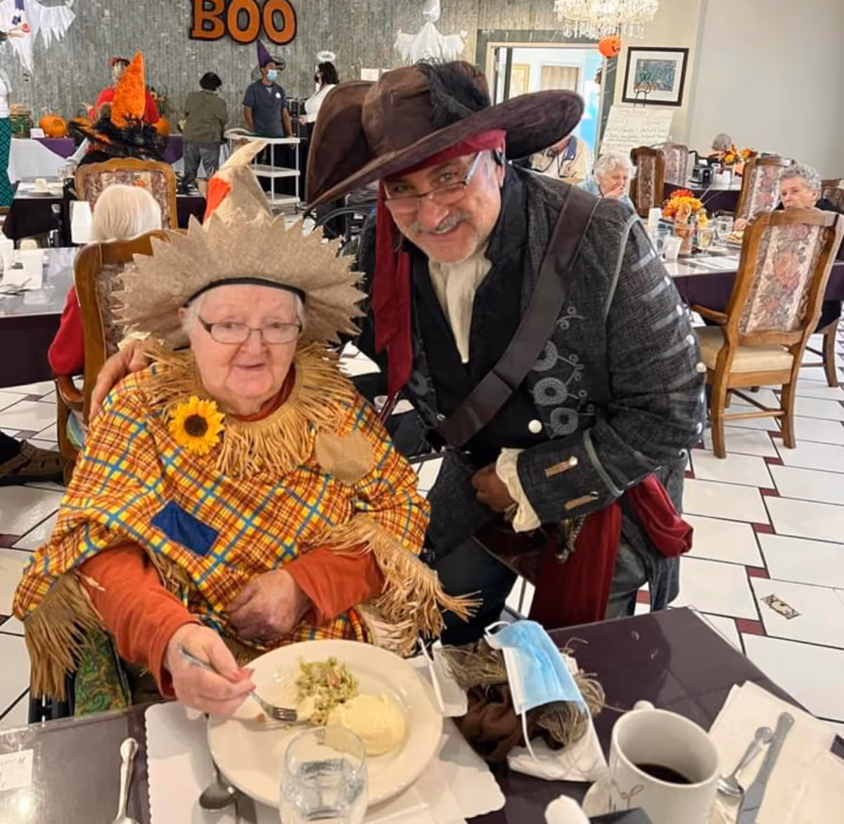 An elderly woman in a scarecrow costume sits at a dining table with a plate while a man in a pirate costume leans in beside her in a decorated dining room.